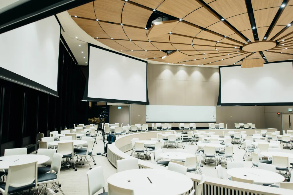 An expansive, well-lit corporate event space, equipped with multiple circular tables each accompanied by sleek white chairs. The area is ready for attendees with pens and notepads set on each table. Overhead, the ceiling boasts an intricate design with wooden panels and circular lighting fixtures. Large blank projection screens at the front await presentations, with a camera setup indicating the potential for live recording or streaming. The setting exudes a professional and sophisticated ambiance, ideal for conferences, seminars, or business gatherings.