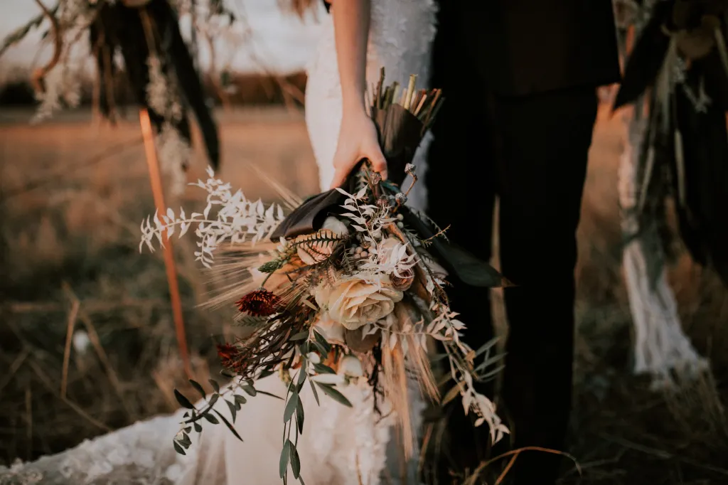 Close-up of a wedding bouquet held by a bride, next to her partner. The bouquet is a stunning collection of flowers including roses and wildflowers, adorned with feathers and greenery, creating a bohemian and earthy aesthetic. The warm, golden hour light bathes the scene, highlighting the intricate details and colors of the bouquet against the soft, romantic backdrop of a field.