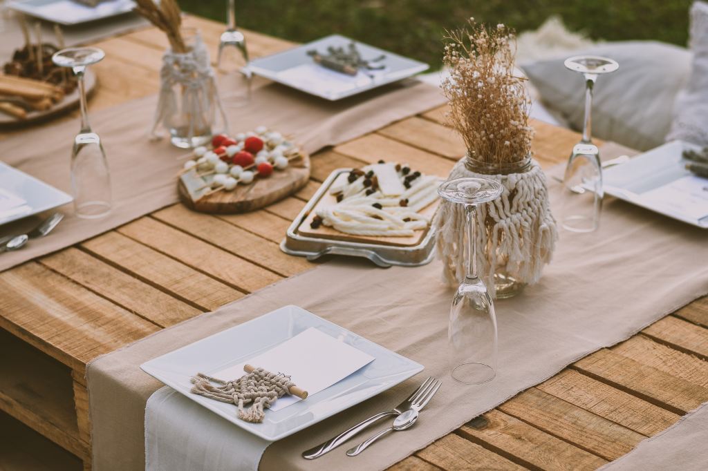 A rustic outdoor party setup featuring a wooden table covered with a natural beige tablecloth. The table is elegantly laid out with square white plates, silver cutlery, and clear wine glasses turned upside down. Decorative touches include a macramé-wrapped glass jar with dried grasses and a wooden board with an assortment of cheese and berries. The setting is informal yet stylish, perfect for a casual social gathering or a laid-back celebration.